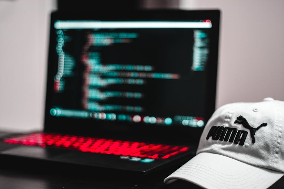 A close-up image of a laptop with a dark screen displaying lines of code in bright teal and red colors, suggesting programming or digital work, placed on a dark surface. In the foreground, a white Pum