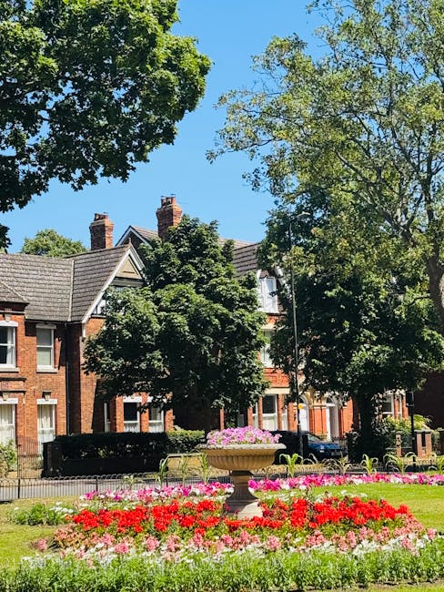 A landscaped urban garden featuring a large, vibrant flower bed in the foreground filled with red, pink, and white blooming flowers, neatly arranged around a classical-style stone planter. Behind the