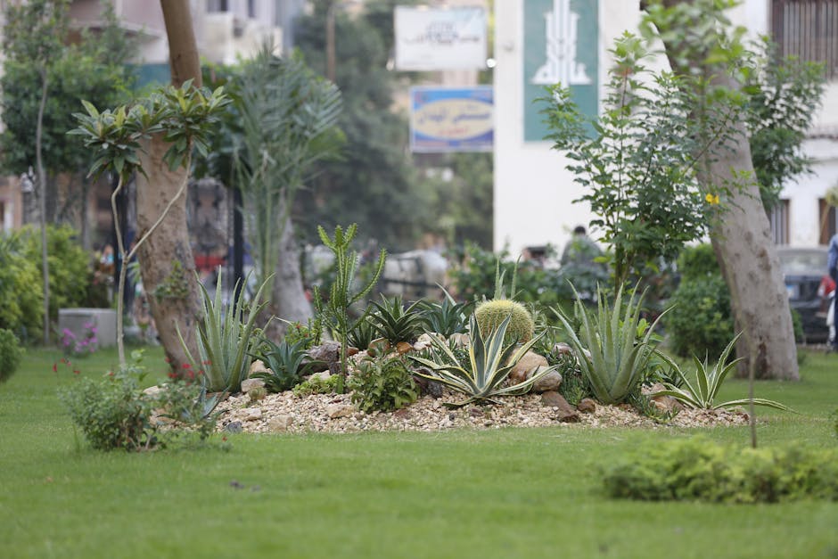A landscaped urban garden featuring a large, vibrant flower bed in the foreground filled with red, pink, and white blooming flowers, neatly arranged around a classical-style stone planter. Behind the